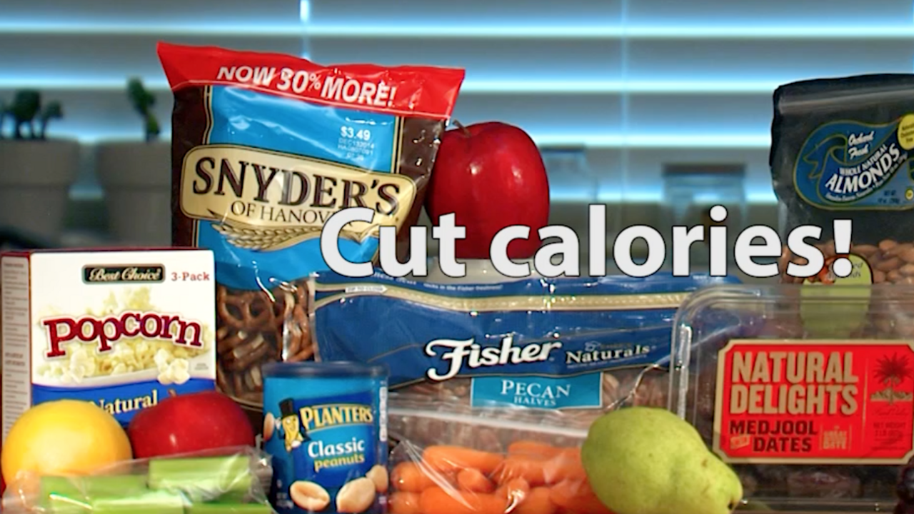 An assortment of snack foods on a butcher block table top, including a green pear, red grapes, baby carrots, pretzels, pecans, dates, almonds, and trail mix. 