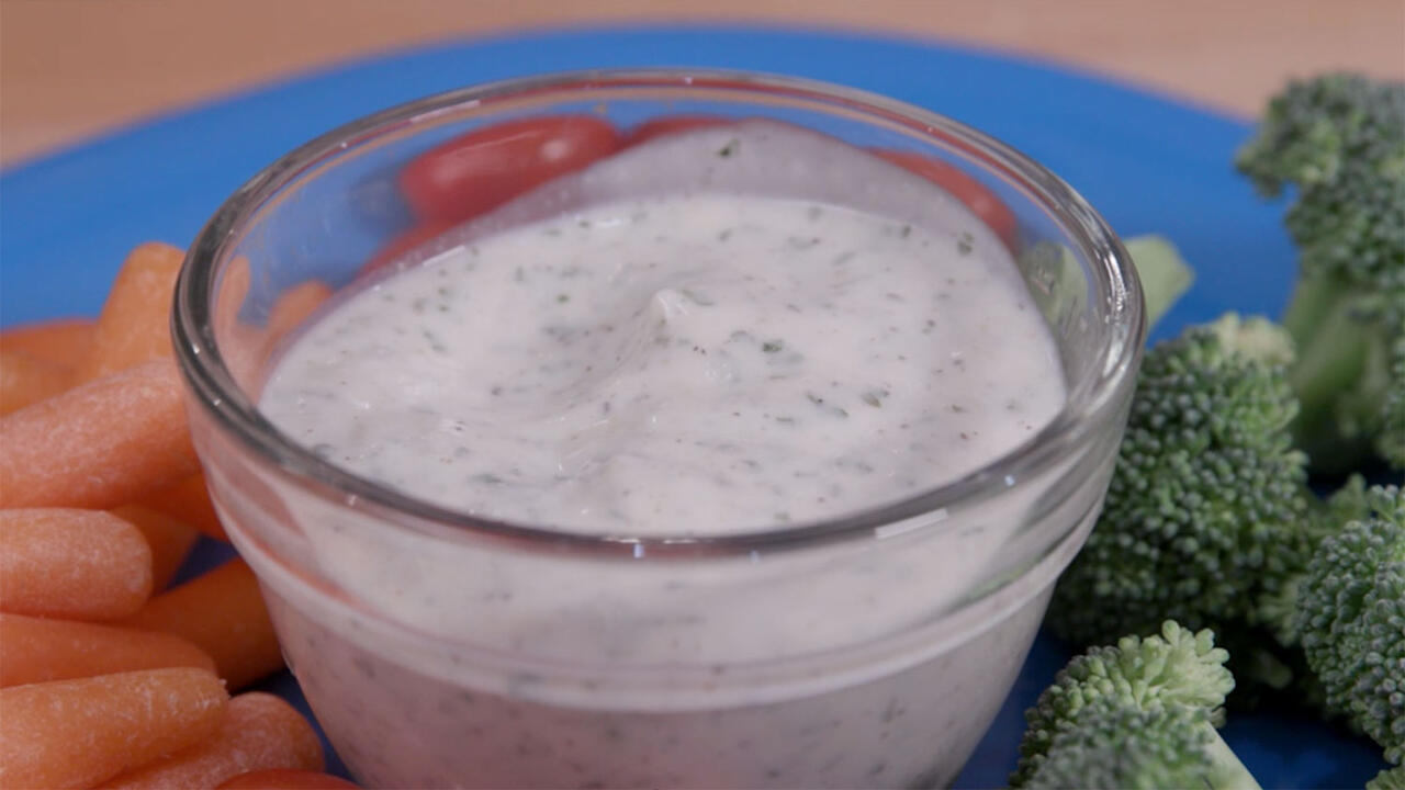 Homemade ranch dressing in a clear glass bowl atop a turquoise blue plate, surrounded by baby carrots, broccoli florets, and grape tomatoes.