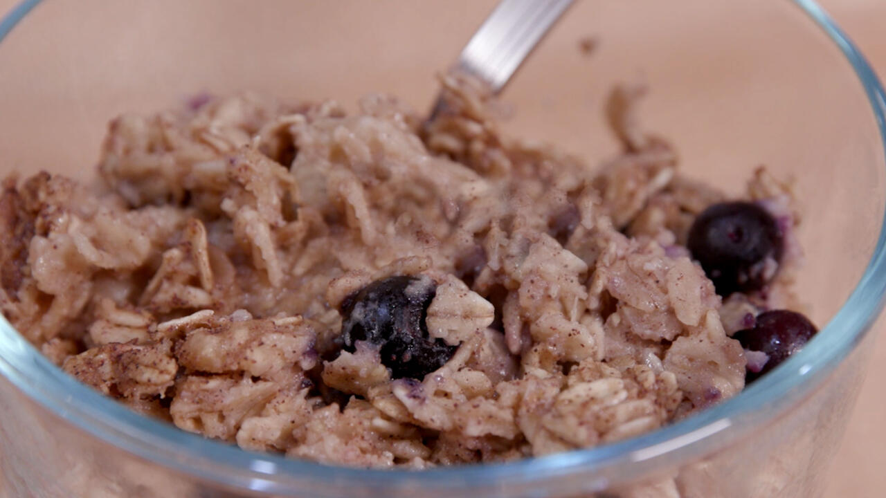 A small clear glass bowl holds baked oatmeal with blueberries and a stainless steel spoon.