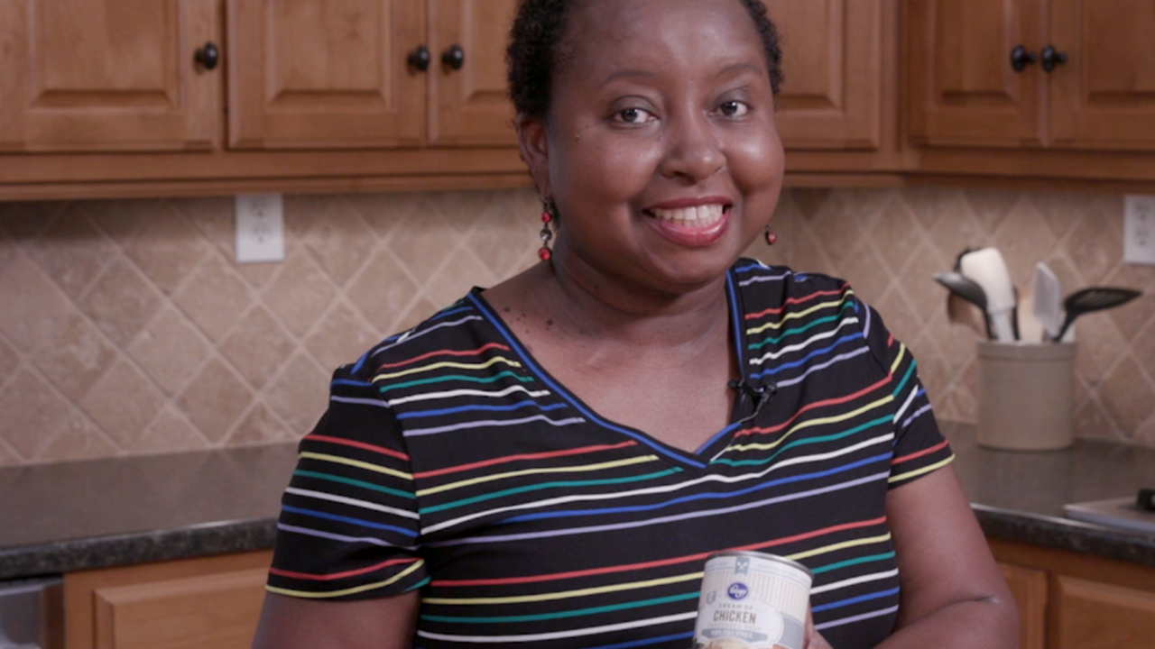 A woman stands in a kitchen while holding a can of reduced fat cream of chicken soup. 
