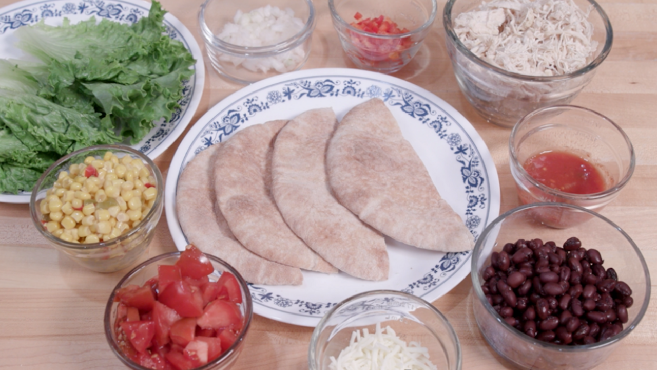 An array of ingredients in glass bowls, including romaine lettuce, onion, diced red pepper, Mexicorn, diced tomato, black beans, salsa, cooked diced chicken breast, and mozzarella cheese, surround a blue and white plate bearing four halves of whole wheat pita bread.