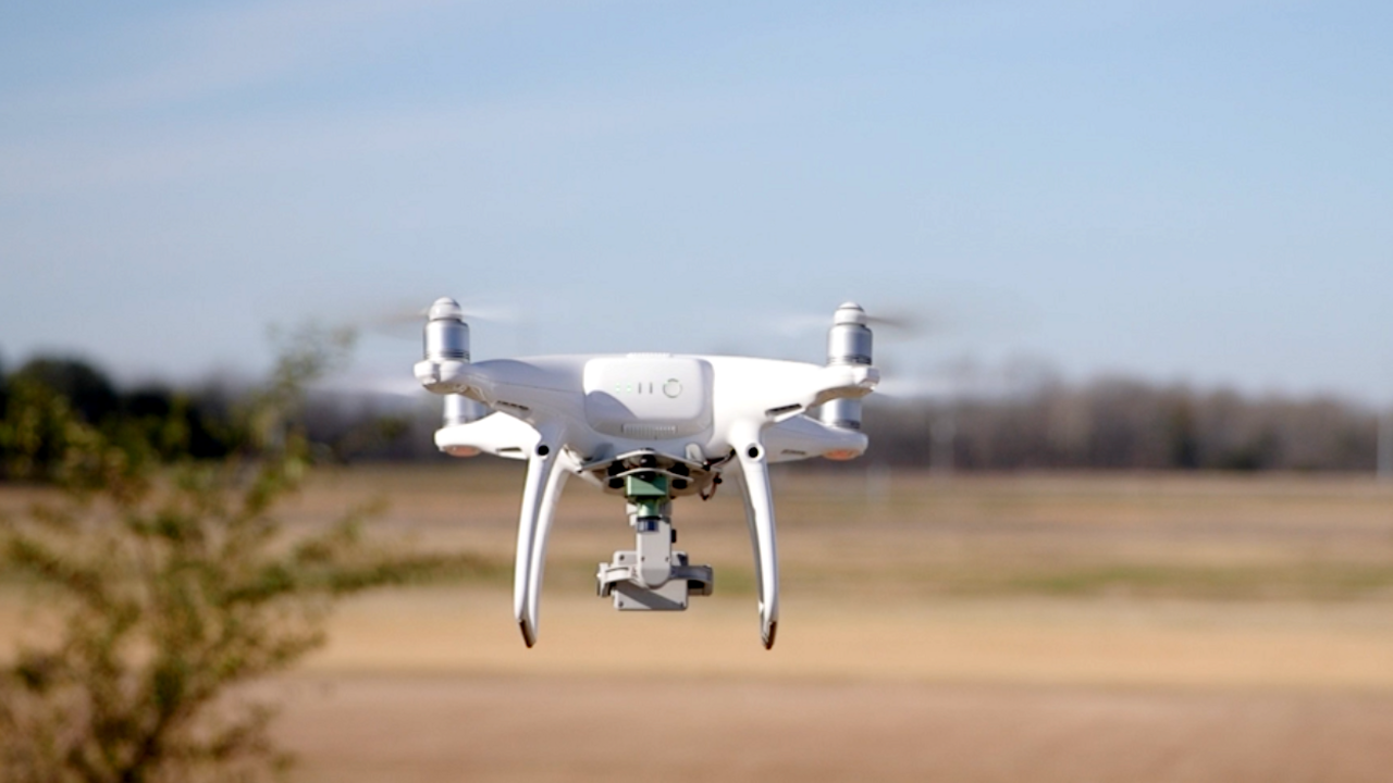 A close up shows a drone hovering above an open field.