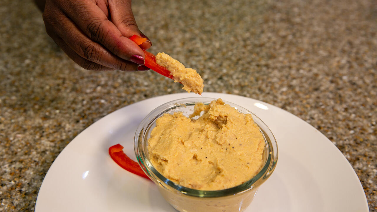 A hand is shown dipping a bell pepper strip into a bowl of garlic chickpea dip.