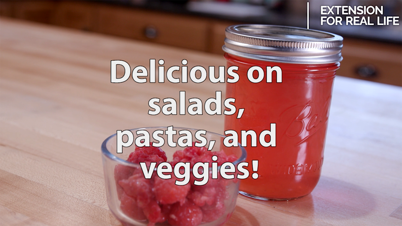 A clear glass bowl of red raspberries sits on a butcher block table in front of a pint clear glass jar filled with homemade raspberry vinegar and topped with a metal lid and ring. Text overlay reads, "Delicious on salads, pastas, and veggies!"