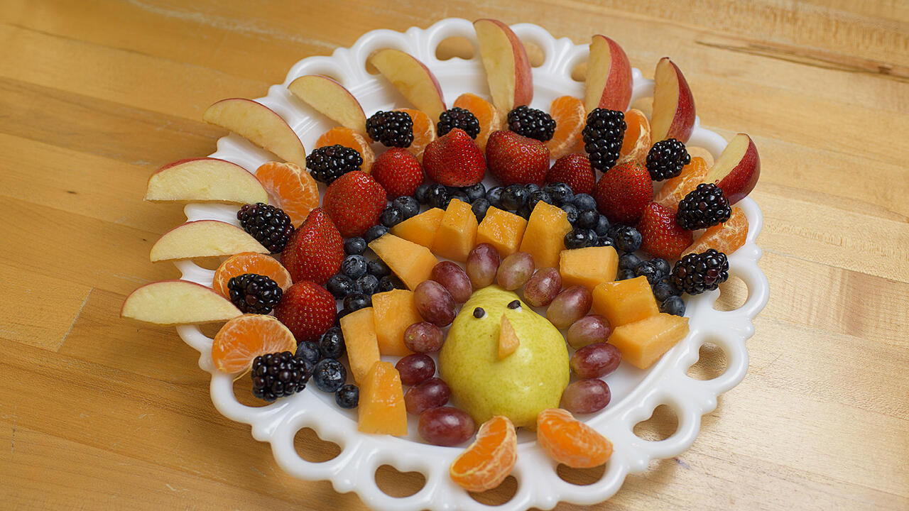 Several rows of fruit including apple slices, tangerine segments, blackberries, blueberries, cantaloupe, strawberries, grapes and a pear are arranged on a white platter in the shape of a turkey.