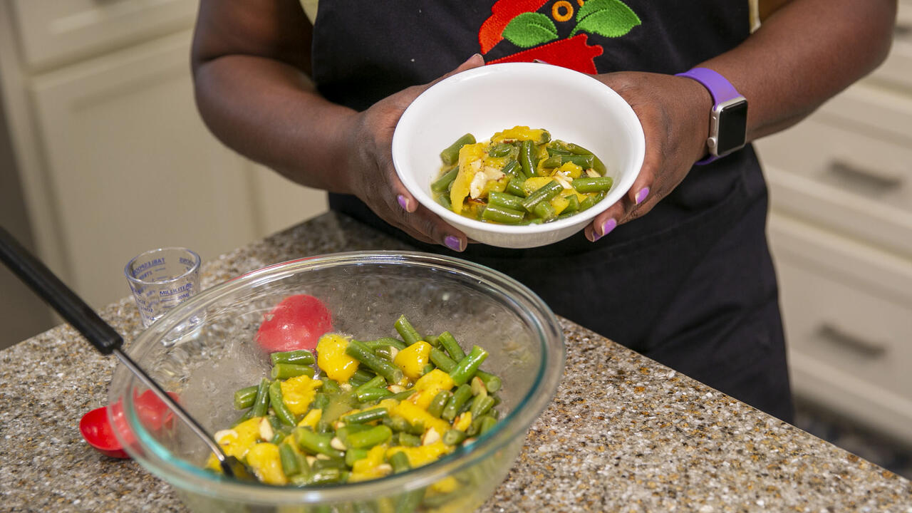 A bowl of peachy green beans sits on the kitchen counter with a woman holding a serving size in a separate bowl.