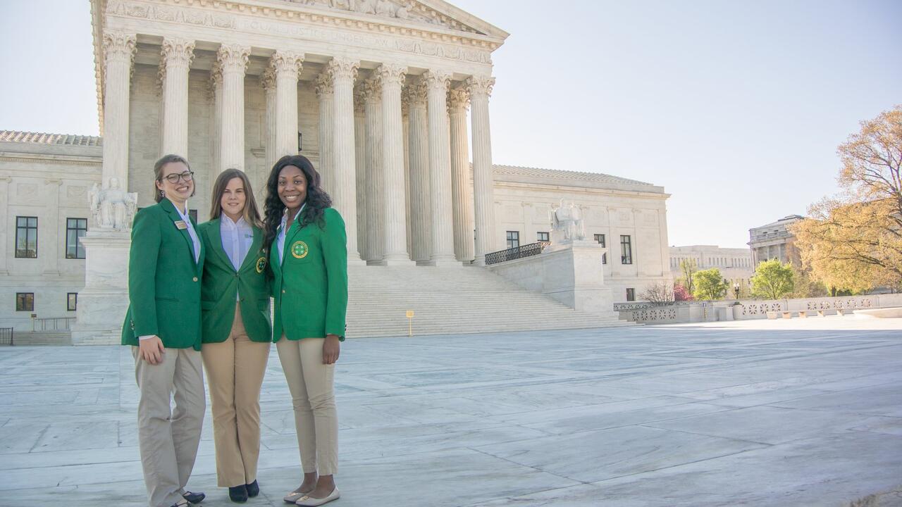 Three female teenagers wearing green blazers and khaki pants standing in front of the Supreme Court Building in Washington, D.C.