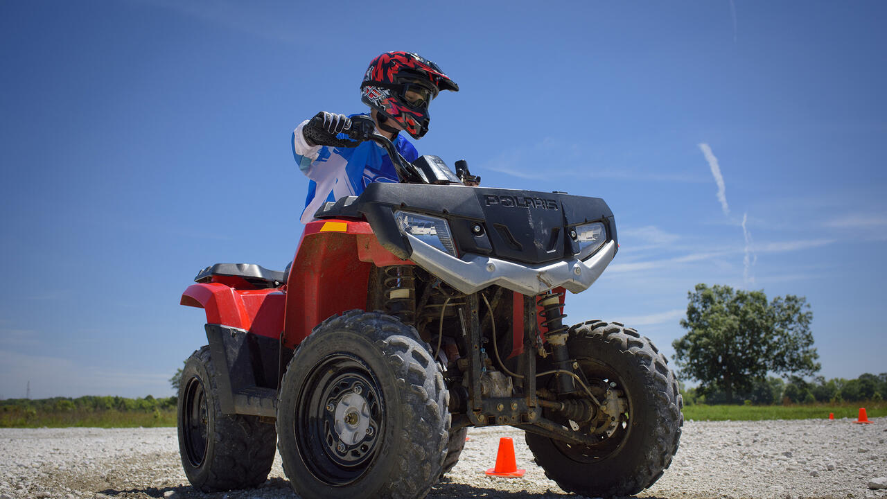 A teenage boy in a blue and white long-sleeved shirt and protective gear, including a red and black helmet and black gloves, rides a red ATV around orange cones during a safety class. (Kevin Hudson)