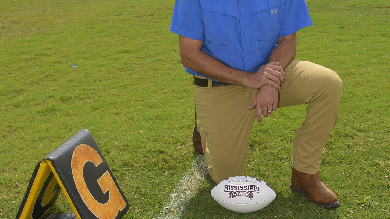A man kneels on a football practice field beside a football.