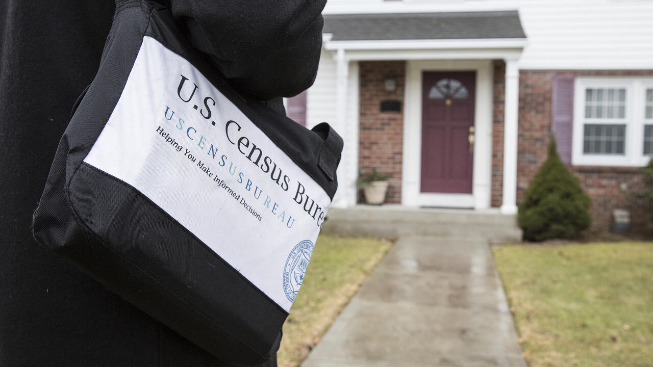 A person carrying a black bag marked with census information approaches the front door of a house.