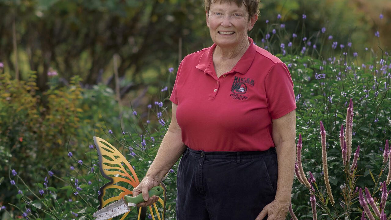 A woman holding a gardening tool stands in a green landscape.