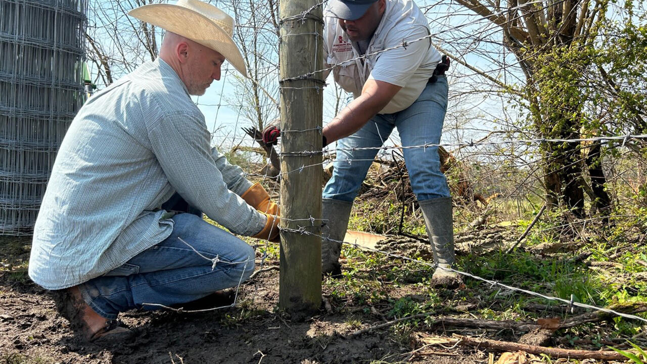 Two men tie barbed wire around a fence post.
