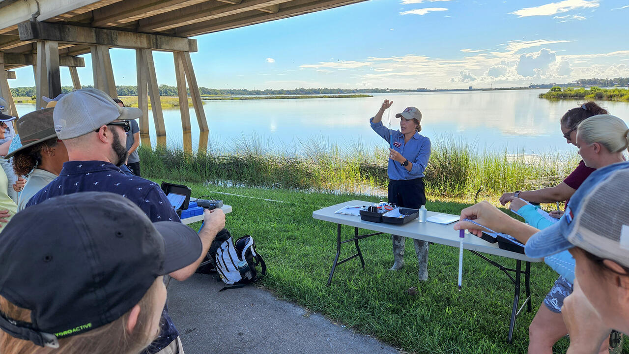 A woman stands at a table under a bridge while others look on.
