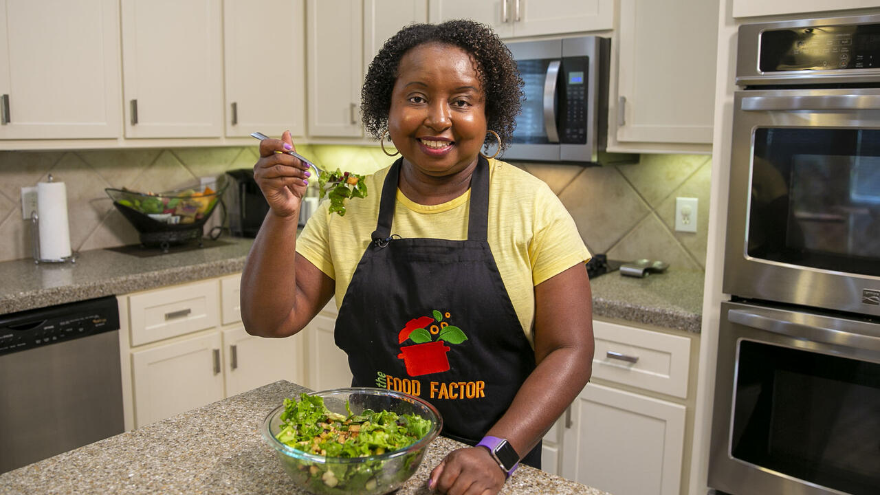 A woman in a kitchen smiles and holds a forkful of salad up for the camera.