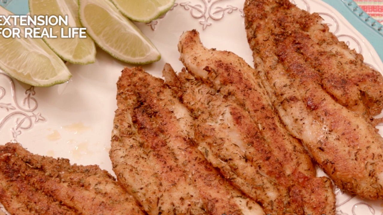 Four browned, seasoned catfish fillets on a white plate garnished with lime wedges.