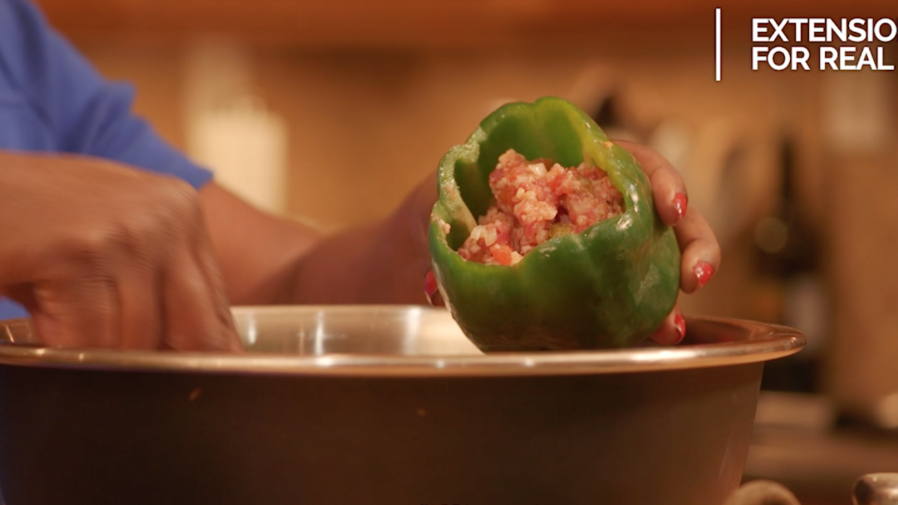 Hands spoon a ground meat and riced cauliflower mixture into a raw green bell pepper held over a stainless steel bowl. 