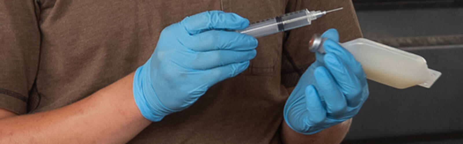 Two gloved hands hold a syringe and a medical container.