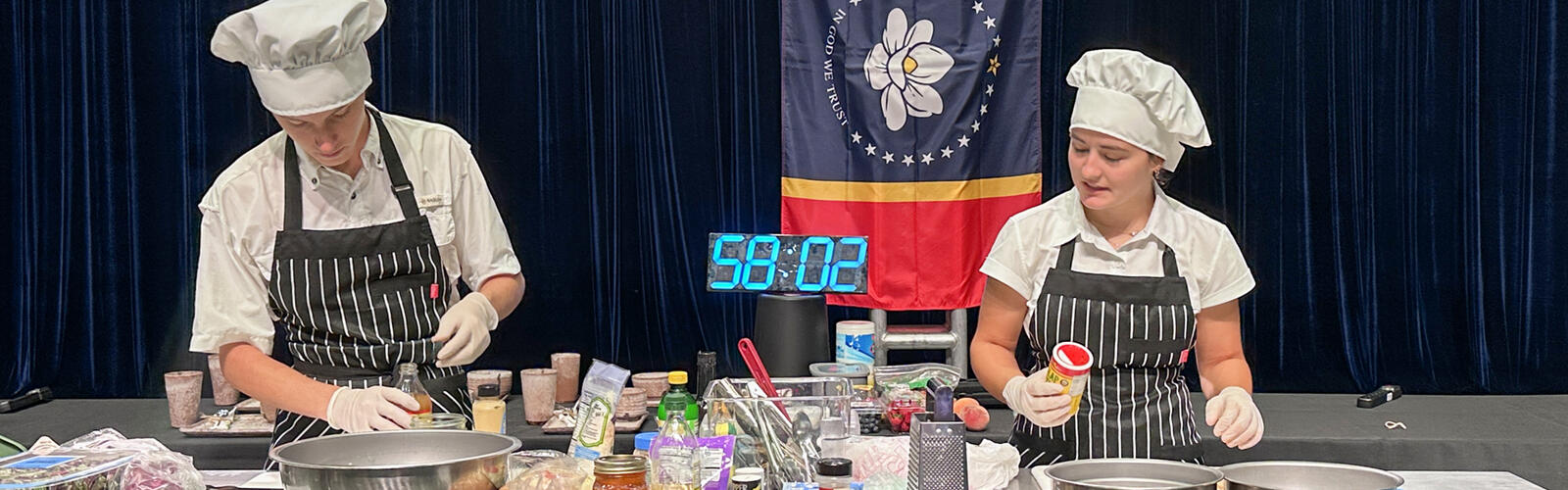 Two people prepare food during a cooking competition with a large timer in the background.