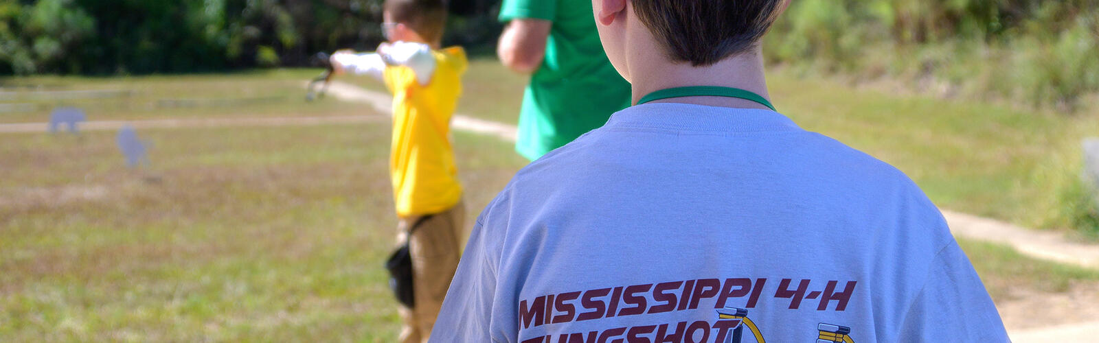 In the foreground, a boy pictured from behind wearing a T-shirt reading “Mississippi 4-H Slingshot 2025 State Championship” looks on as another boy aims at a target with a slingshot.