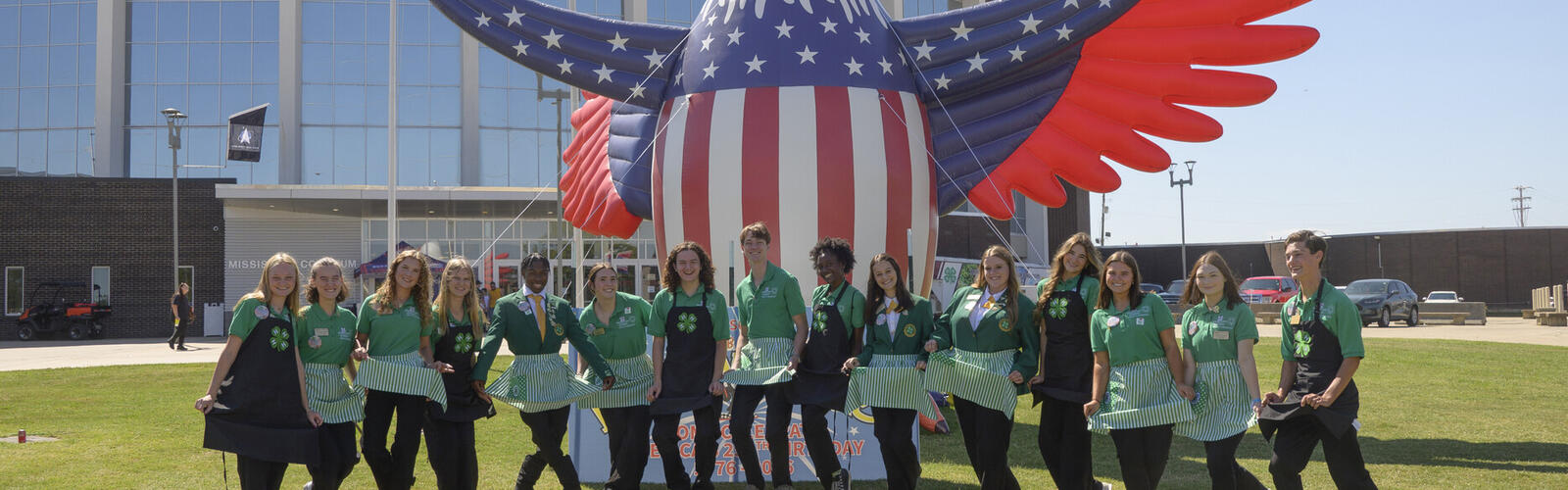 Fifteen young people wearing green jackets stand in front of an inflatable eagle.