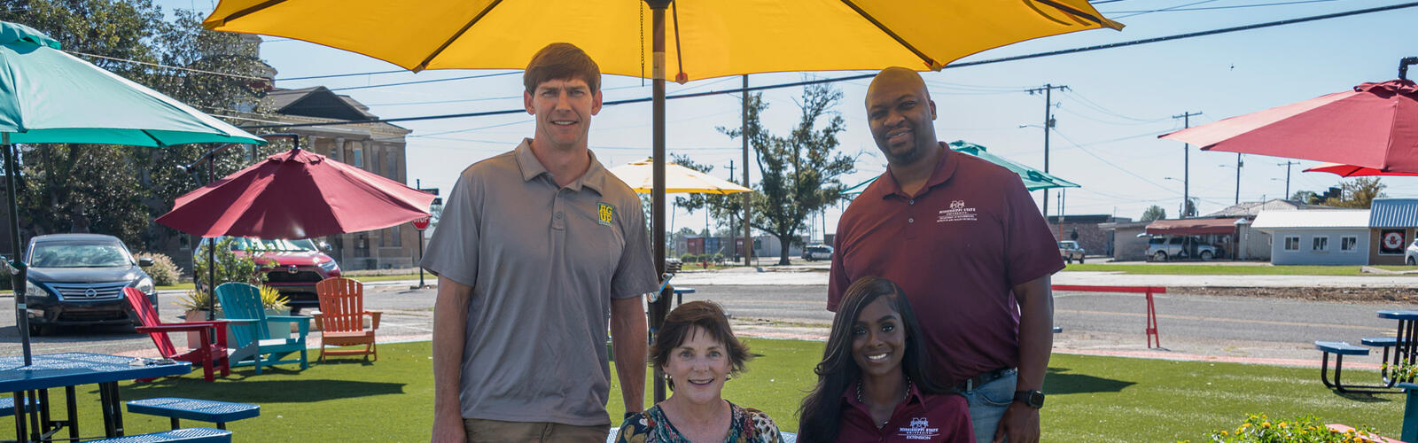 Two people standing and two people sitting under a yellow parasol.