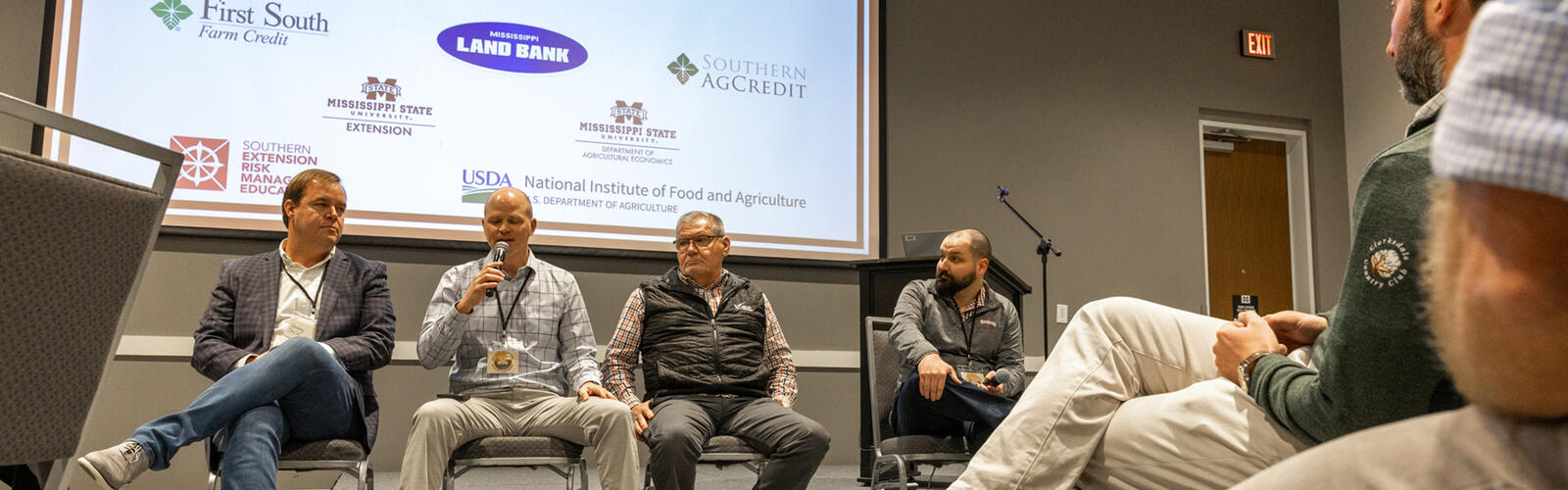 A panel of four speakers sits on a stage in front of a large presentation screen while attendees seated in the foreground listen. The screen displays the conference title, dates, and logos of participating organizations.