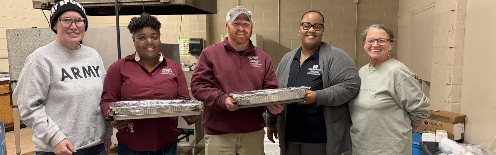 A group of five people standing together in an industrial-style room, with two of them holding large foil-covered food pans. Boxes, supplies, and equipment are visible around the room.
