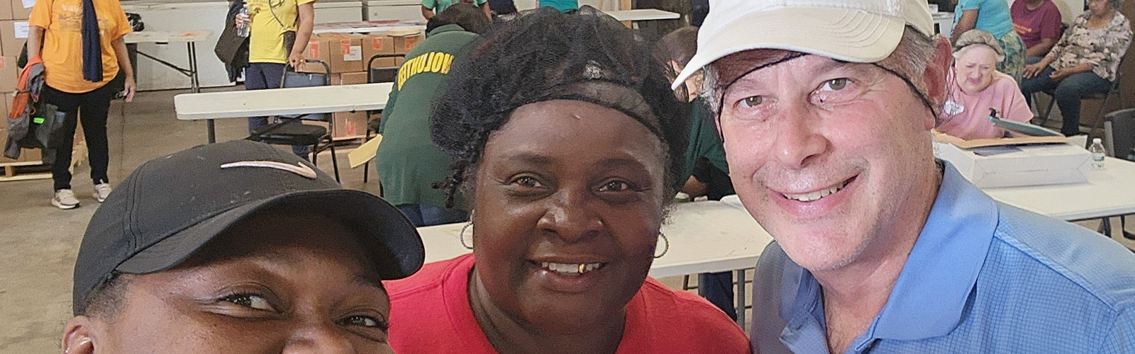 Three people pose for the camera in a church auditorium with others in the background as they prepare to distribute food.