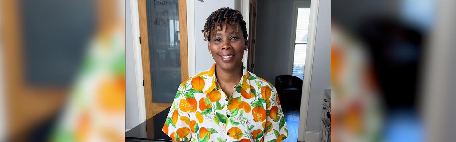 A woman stands in a kitchen with a cutting board, kitchen scissors, a vegetable peeler, and measuring cups on the counter in front of her.