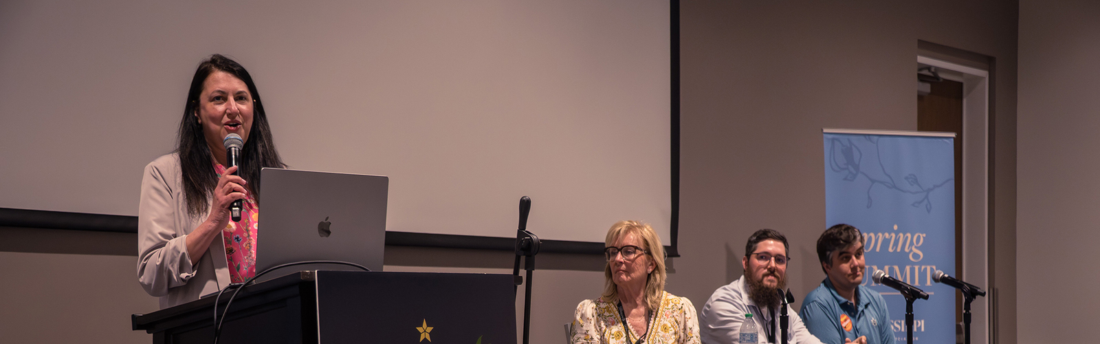 A speaker at a Mississippi Tourism Association podium addresses a panel seated behind a table in an auditorium.