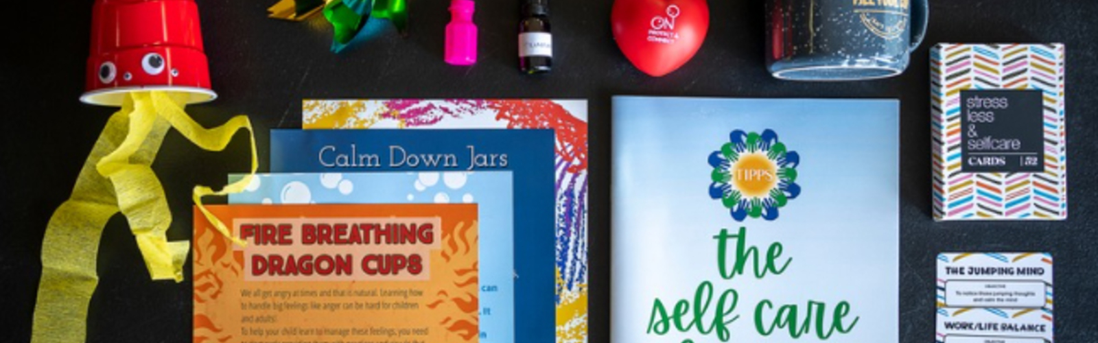 The contents of one toolkit, including a homemade fire-breathing dragon cup, various literature on parental self-care, strategies for emotion regulation and directions for crafts that support hand-eye coordination and fine motor skills, along with a coffee mug, essential oils, handheld pinwheel, a stress ball and bubbles. 