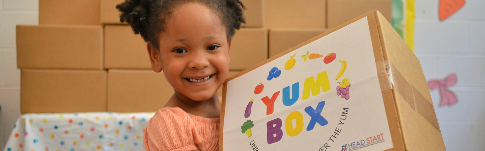A young girl smiles while holding a colorful “Yum Box” package from Head Start, with stacked cardboard boxes and a “Welcome Spring” bulletin board visible behind her.