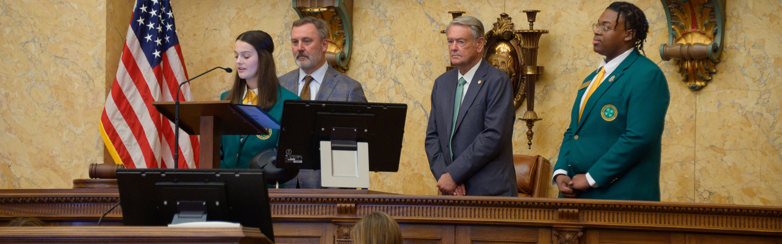 A teenager speaks at a podium as two adults and another teen look on.