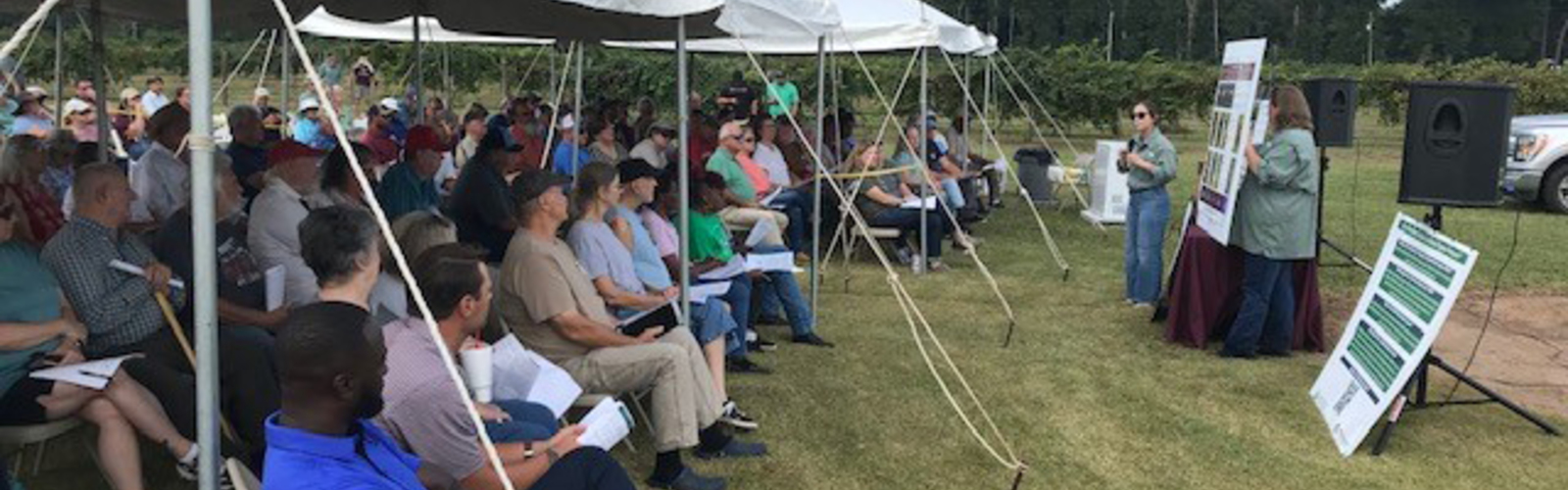 Presenters address a large crowd sitting under outdoor tents.