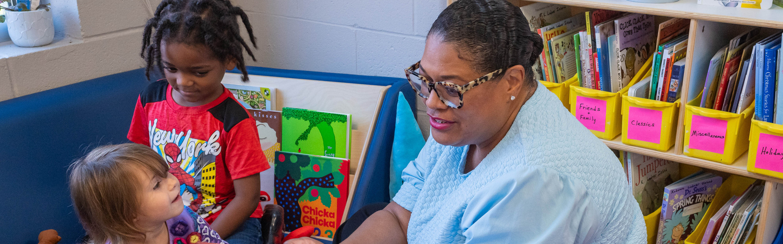 An adult reads from a picture book to two young children seated in a classroom reading corner.
