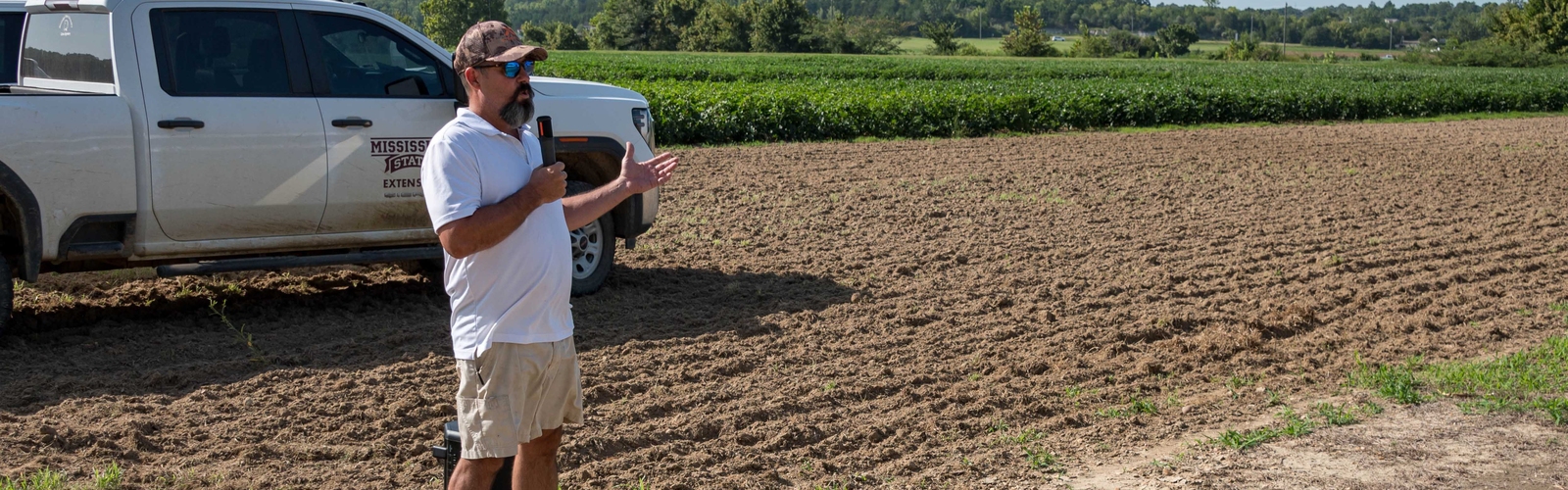 A speaker stands in front of research plots with a Mississippi State University Extension pickup truck in the background.