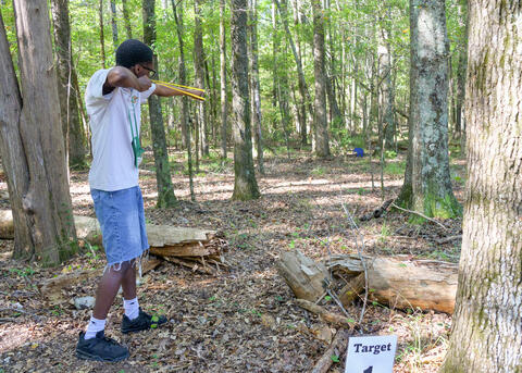 Teenager in a wooded area drawing back the pouch of a slingshot aimed at a target.