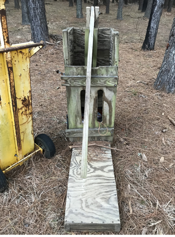 A man-made hand baler made of wood sits on pine needle-covered ground in a forest.