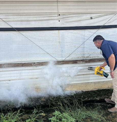 A person holds a smoke generator outside of a building. Smoke is being directed toward the bottom of the wall.