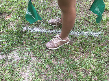 Two feet on opposite sides of a white chalk line on the ground, with two green wire flags in the ground, one in front of the left foot and one behind it.