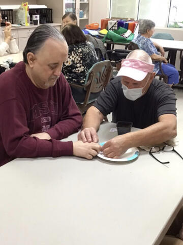 Two people sit at a table; one is helping the other to place a tomato seed on a paper plate.
