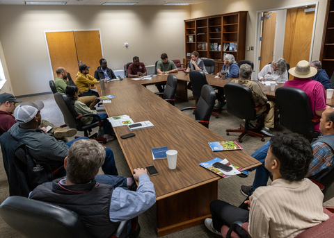 A group of people sit around tables in a meeting.