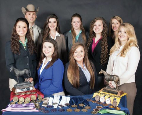 Several people pose behind a table full of medals and two horse trophies.