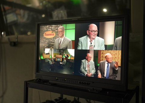 Video monitor displays a talk show set with two men in suits being interviewed, shown from multiple camera angles, with a “Farmweek” sign in the studio background.