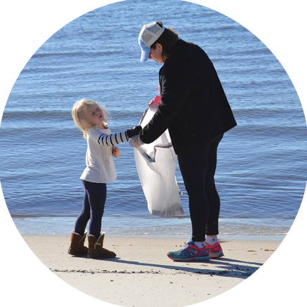 An adult and child standing on a beach put debris into a trash bag.