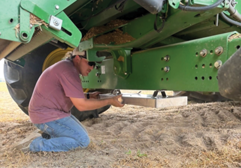 A person is kneeling while mounting a drop pan under a combine.