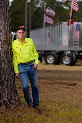 A man wearing a bright yellow button-up shirt leans against a large truck and rests one hand in his blue jean pocket.