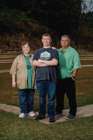 A teenage boy wearing a Shooting Sports National Championships shirt stands between his mother and father as they all smile.