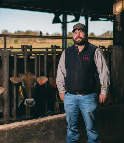 A man gazes off to the left as the sun sets behind him and two cows look at him from the opposite side of the fence he is leaning on.