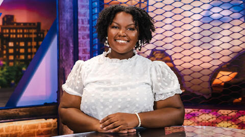 A smiling woman wearing a white shirt sits behind a desk with her hands resting on the desk in front of her.